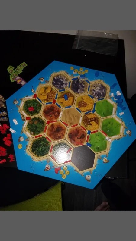 Picture of a catan board - 19 interlocking hexagons with natural terrain art, forming an island. A series of red and blue blocks form roadways between the hexes.