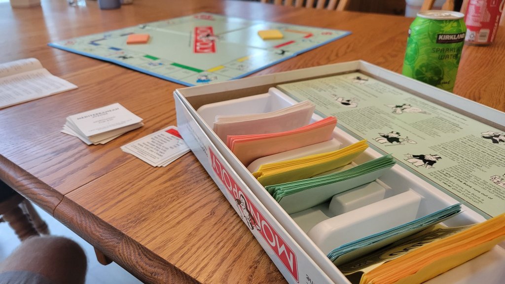 An open monopoly board on a wooden table, mid game. Cards and pieces are laid across the table